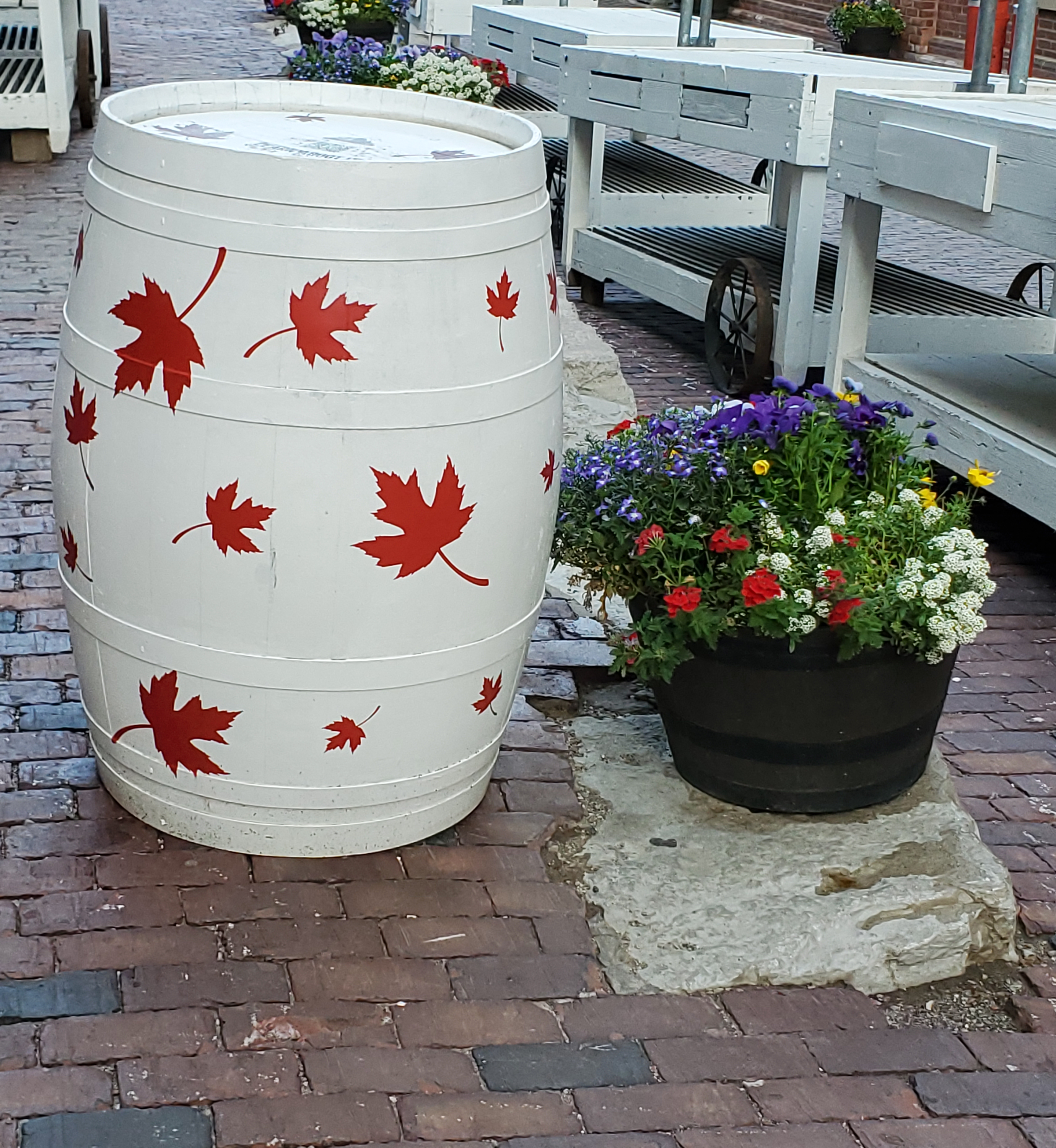 A white barrel with maple leaves next to a bouquet of flowers on a brick road.